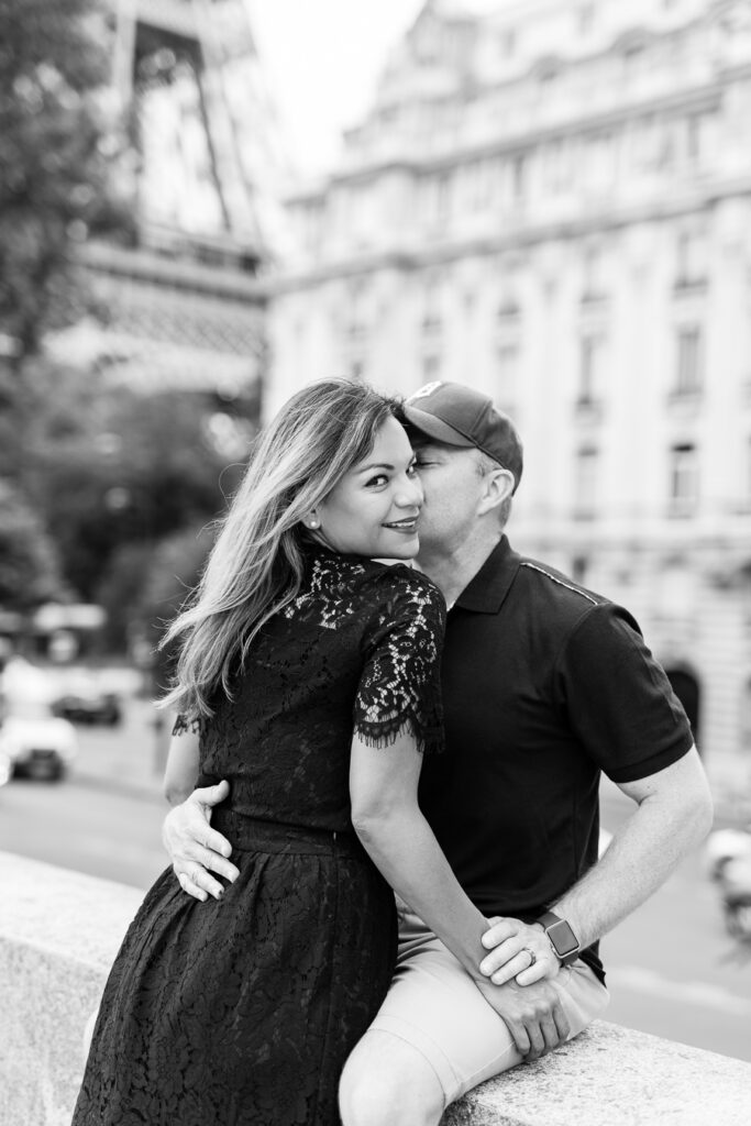 Black and white photo of a couple laughing against the wall of the Pont Neuf, Paris rooftops behind