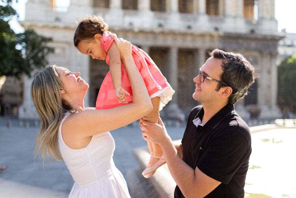 Mom lifting her laughing daughter into the air while Dad watches, in a Paris courtyard