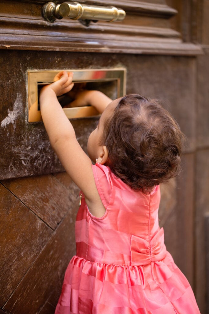 Little girl in a pink dress reaching up to a brass mail slot on a wooden Parisian door