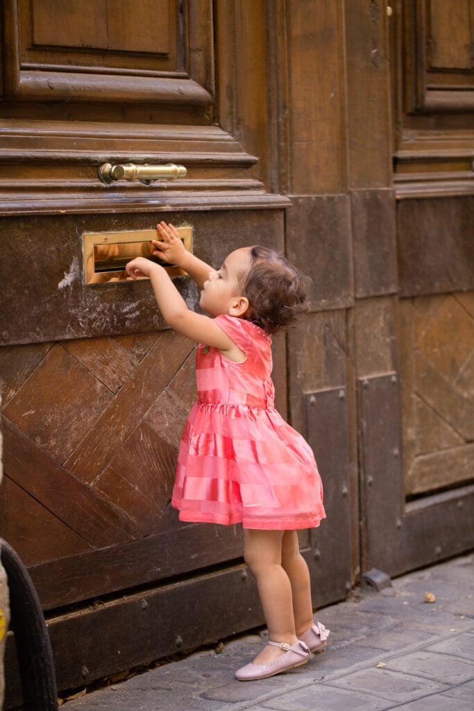 Little girl in a pink dress standing on tiptoe at a large wooden door in Paris