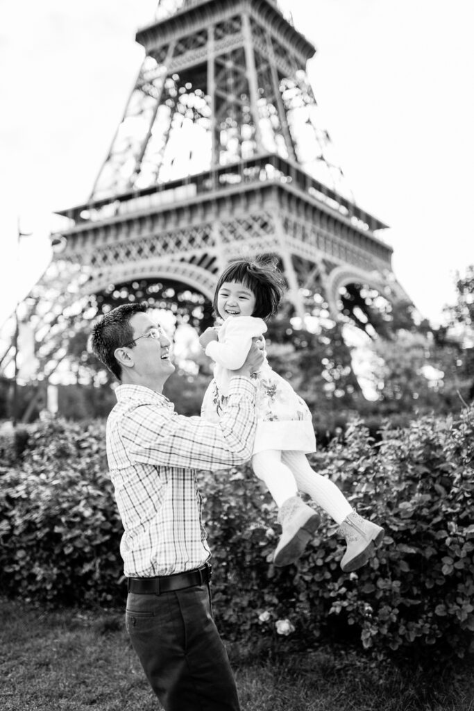 Black and white photo of Dad lifting his laughing daughter in front of the Eiffel Tower