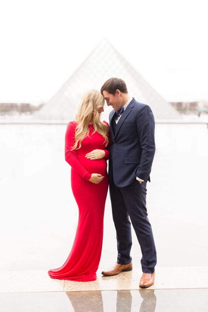 Expecting couple embracing in front of the Louvre pyramid, mom-to-be in a long red dress