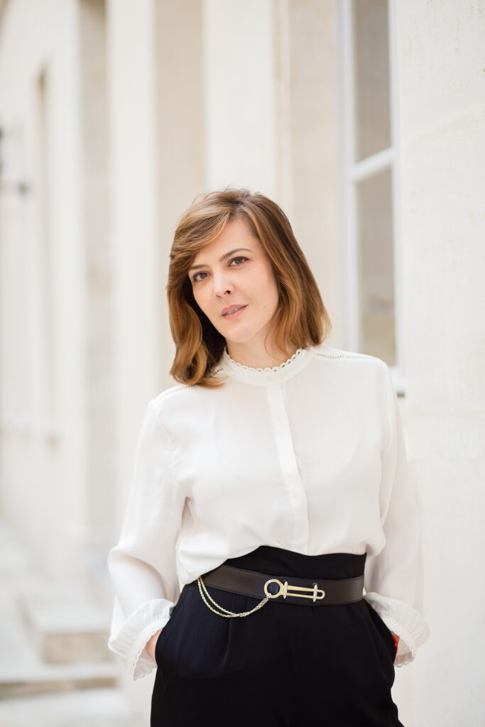Solo portrait of a woman in a white blouse and black trousers in front of pale Parisian shutters