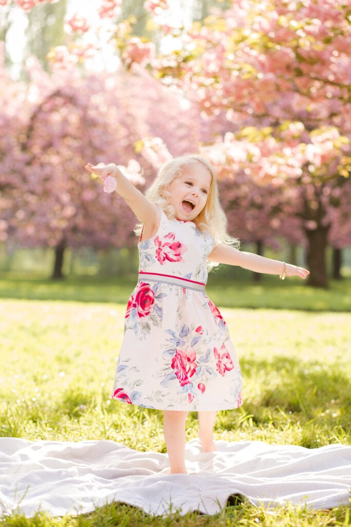 Little girl in a floral dress throwing her arms wide under the cherry blossoms at Parc de Sceaux