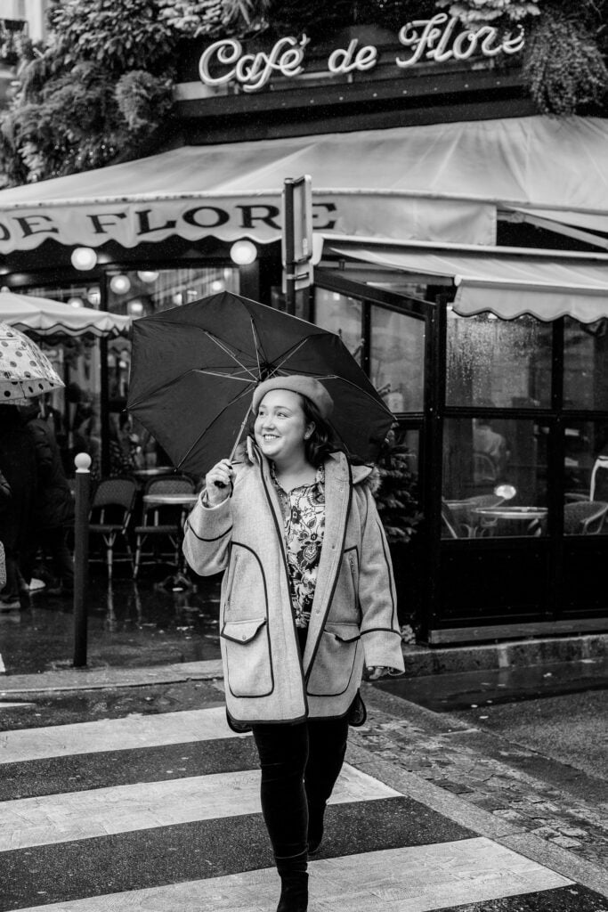 Black and white portrait of woman walking across street in Paris on rainy day with umbrella by Katie Donnelly Photography