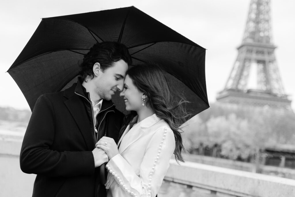 Black and white portrait of couple embracing under umbrella in front of Eiffel Tower by Katie Donnelly Photography
