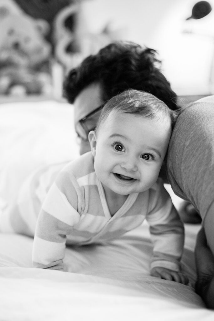 Black and white photo of a smiling baby with bright catchlights in her eyes as Dad kisses her head