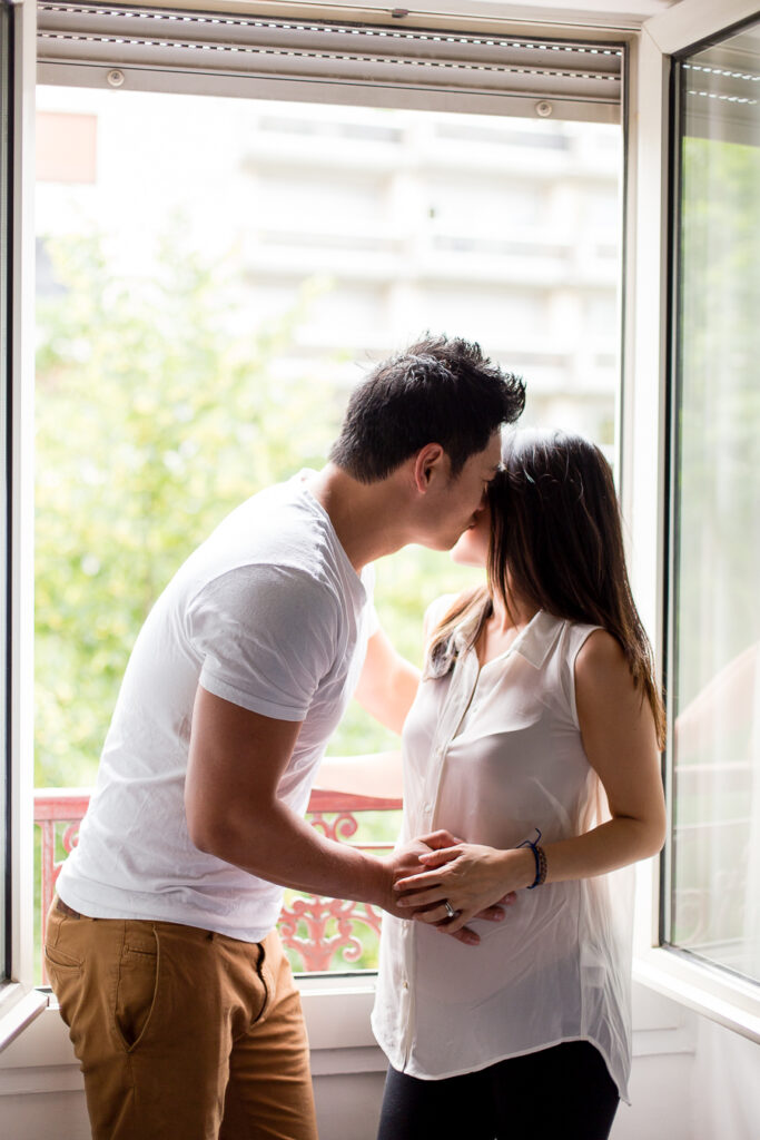 Expecting couple kissing by an open window in soft natural light