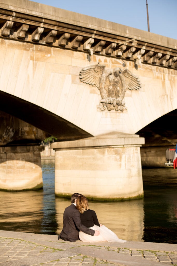 Couple sitting together on the bank of the Seine beneath a stone bridge in Paris