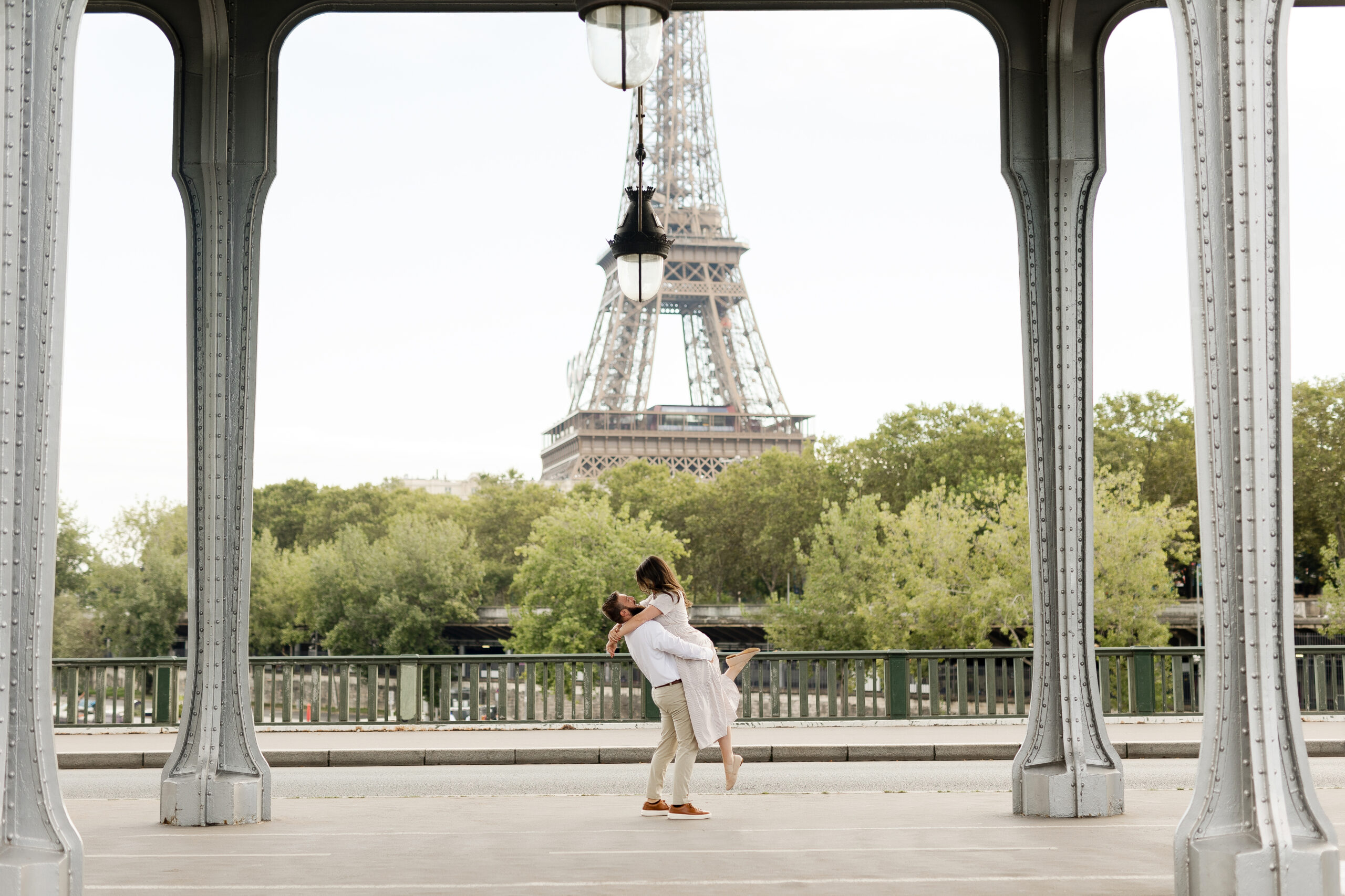 A romantic couple photoshoot on Bir Hakeim Bridge in Paris, captured by Katie Donnelly Photography. The couple is posing under the iconic bridge with the Eiffel Tower in the background.