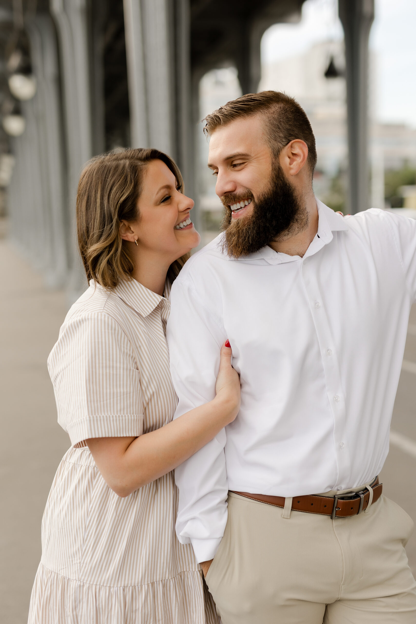 A romantic couple photoshoot on Bir Hakeim Bridge in Paris, captured by Katie Donnelly Photography. The couple is posing under the iconic bridge with the Eiffel Tower in the background.