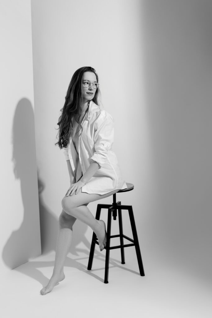 Woman in an open white shirt seated on a stool in a bright Paris boudoir studio, in black and white