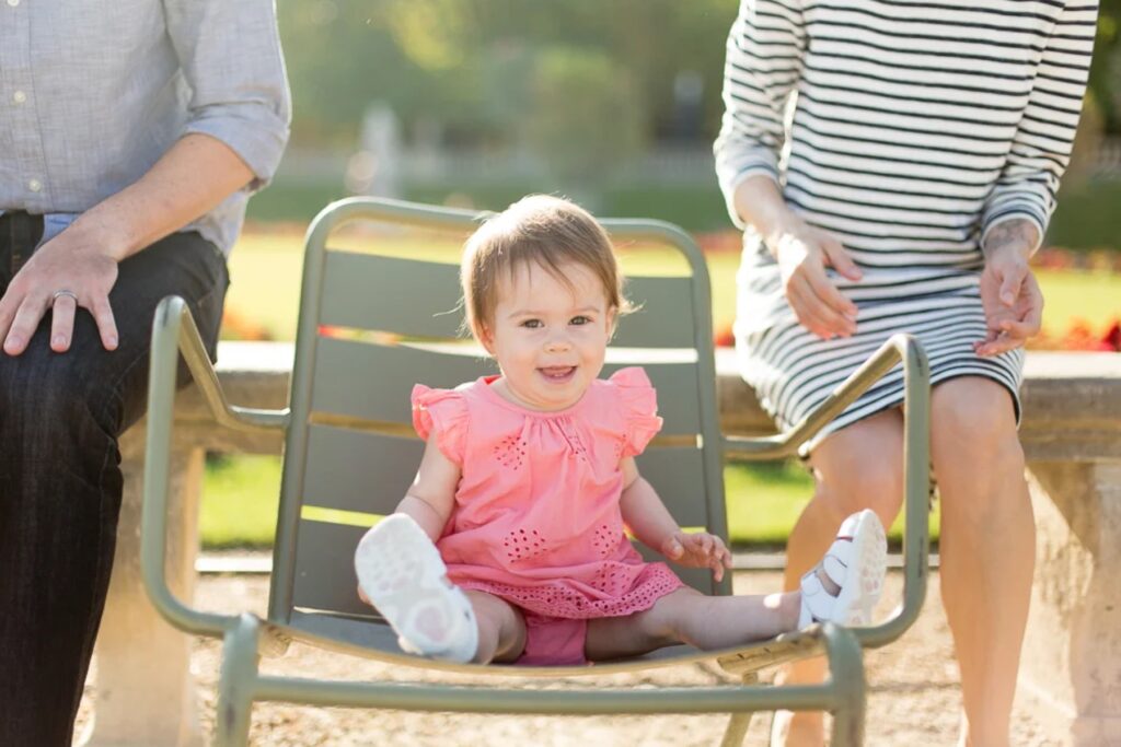 Baby girl in a pink romper sitting on a green garden chair between her parents