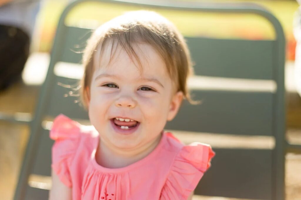 Close-up of a smiling baby girl in a pink romper on a green chair in a Paris garden