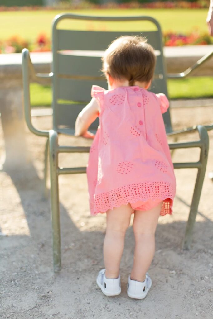 Baby girl in a pink romper standing on a green chair in the Luxembourg Gardens
