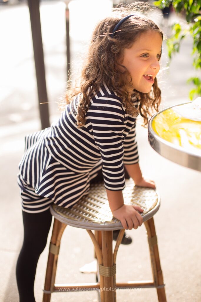 Little girl in a striped top climbing onto a café stool in morning light in Paris
