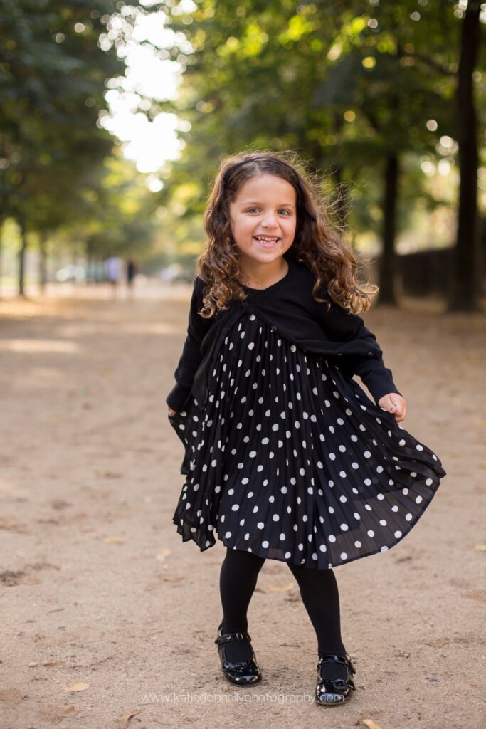 Little girl twirling in a polka dot dress on a tree-lined path in a Paris park