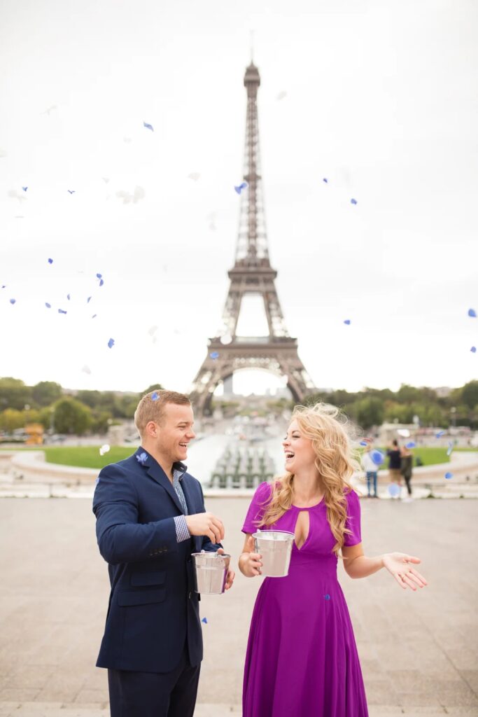 Couple tossing blue confetti petals for a baby gender reveal in front of the Eiffel Tower at Trocadéro