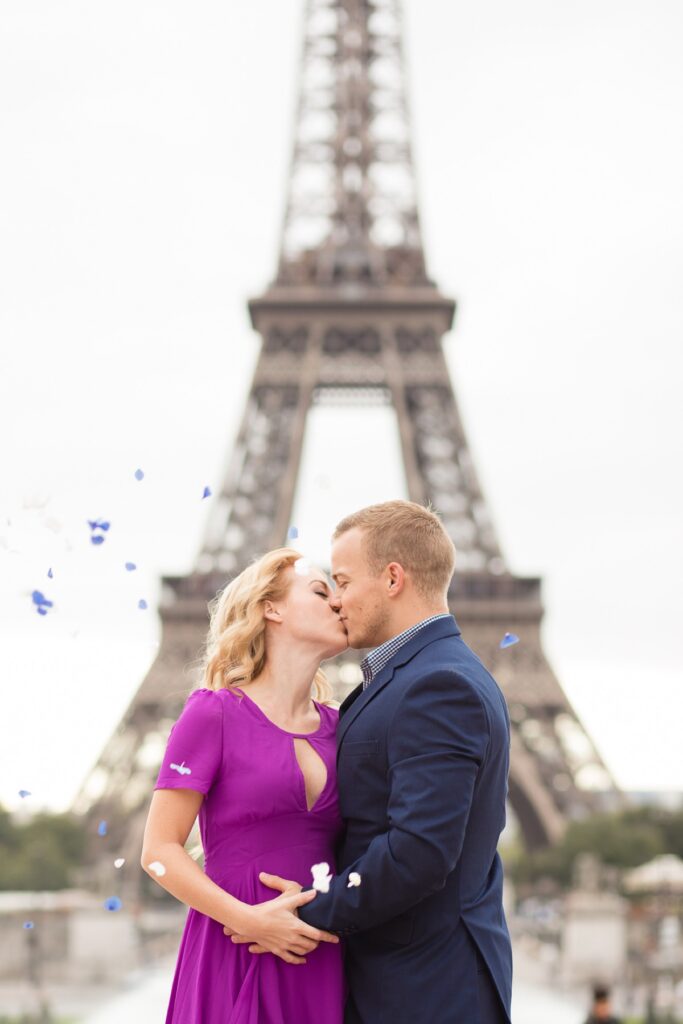 Expecting couple kissing as blue petals fall in front of the Eiffel Tower in Paris