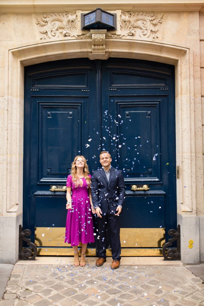 Expecting couple throwing blue petals in front of a navy blue Parisian door for a gender reveal