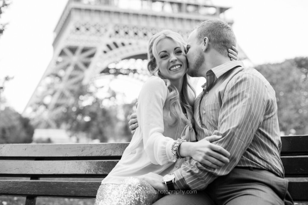 Black and white photo of a couple cuddling on a bench beneath the Eiffel Tower