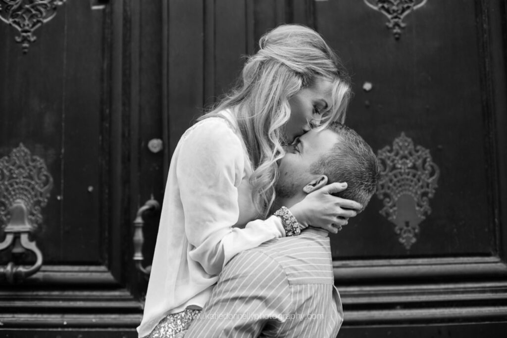 Black and white photo of a couple kissing in front of an ornate Parisian door