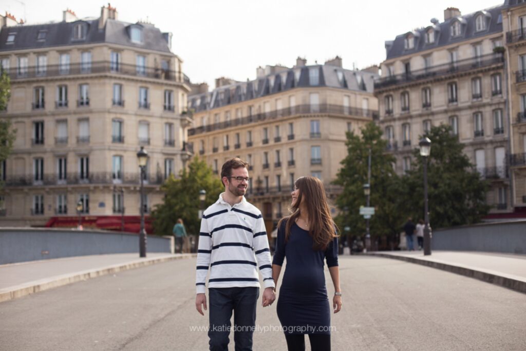 Expecting couple walking hand in hand across a Paris square lined with Haussmann buildings