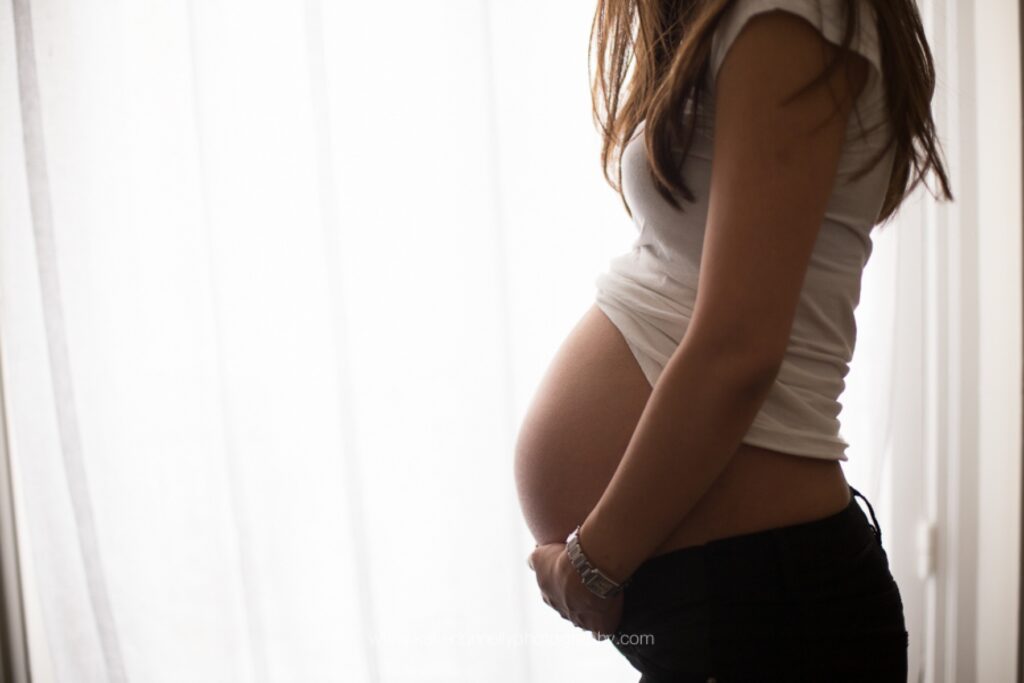 Silhouette of a pregnant woman in profile beside a bright window with sheer curtains