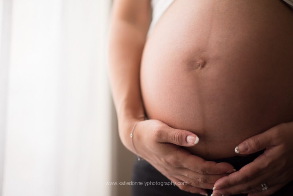 Close-up of a mom-to-be's hands cradling her bare baby bump in window light