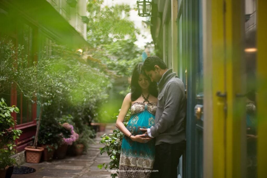 Expecting couple cuddling under the greenery of a covered passage in the Marais