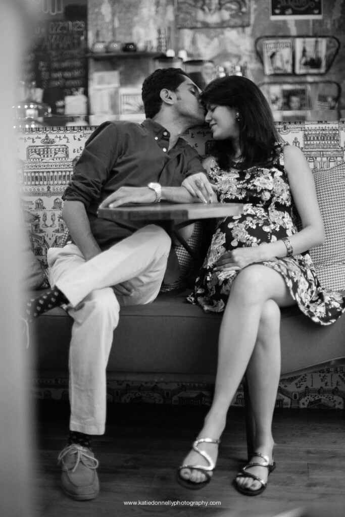Black and white photo of an expecting couple kissing on a sofa in a Paris coffee shop