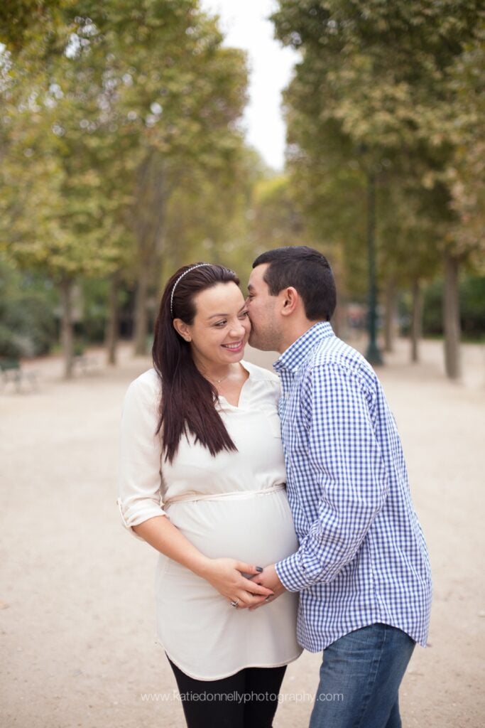 Expecting couple kissing on a tree-lined path in a Paris garden