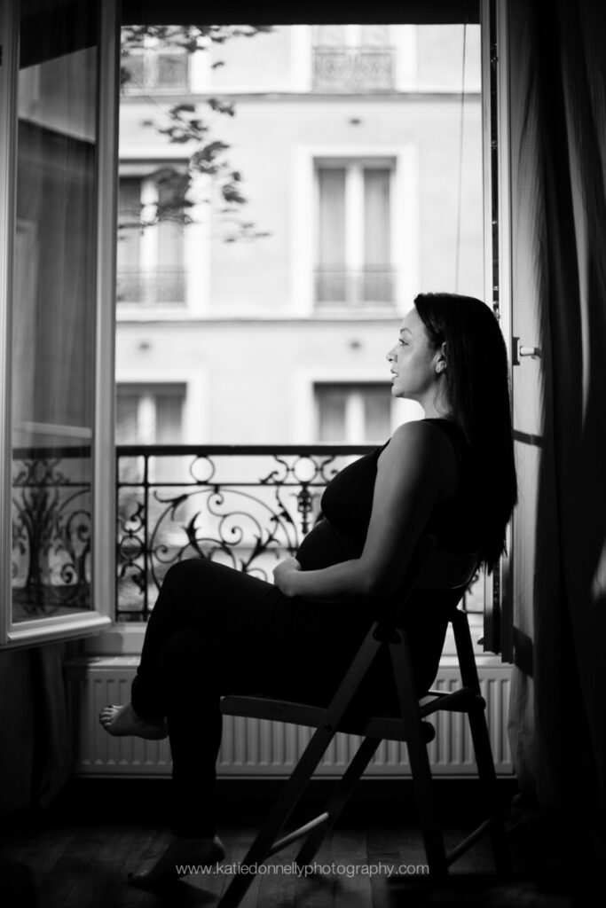 Black and white silhouette of a pregnant woman sitting at an open window with a wrought iron balcony