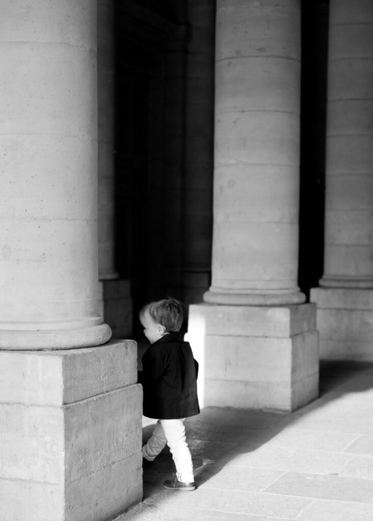Black and white photo of a toddler peeking around a stone column at the Palais Royal