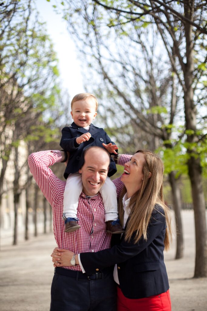 Toddler on Dad's shoulders as his parents laugh in the Palais Royal gardens