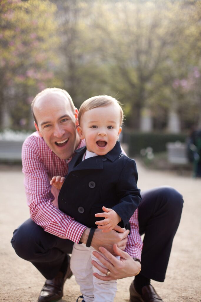 Dad crouching to hug his laughing toddler son in the Palais Royal gardens
