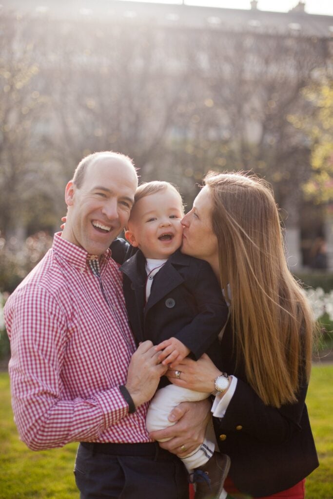 Parents kissing their giggling toddler son in the Palais Royal gardens